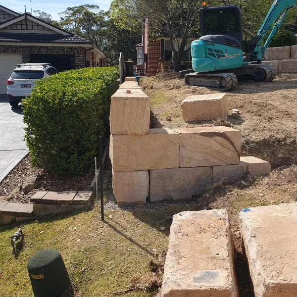 Sandstone block retaining wall construction in Western Sydney with excavator preparing residential sloped yard by West Sydney Landscapes.